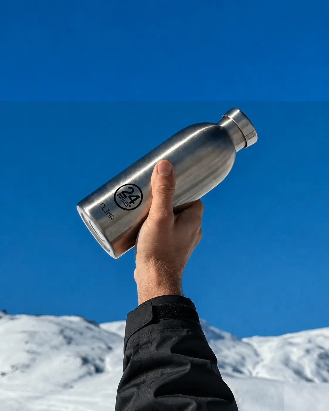 A hand holding a steel 24Bottles Clima against blue sky and snowy mountains.